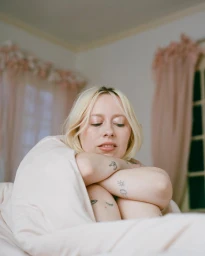Portrait of a blonde woman wrapped in a blanket with visible tattoos on her arms and knees in a bedroom setting