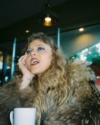 Woman in a furry brown coat with curly hair and blue eyeshadow holding her hand to her face beside a white coffee mug.