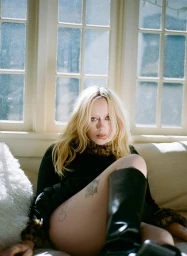 Blonde woman wearing black top and tall black boots seated on a sofa indoors with large multi-pane windows.