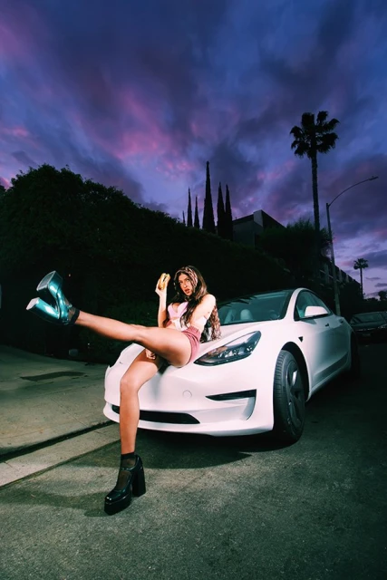 Young woman in high heels sits on a modern white car holding a burger against a city street and sky with palms and plants.
