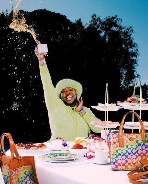 Adult male in lime green sweater pours tea at outdoor tea party with colorful handbags and macarons in a backyard setting.