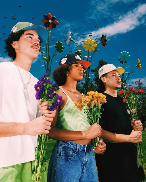 Three people stand in a field of flowers wearing branded baseball caps, holding bouquets under a bright, slightly hazy sky.