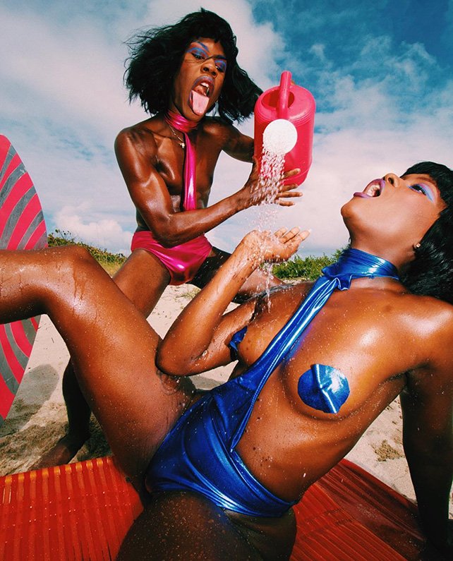 Two dark-skinned adults, one pouring water from a pink watering can, wearing glossy blue and pink outfits on a sunny beach scene.