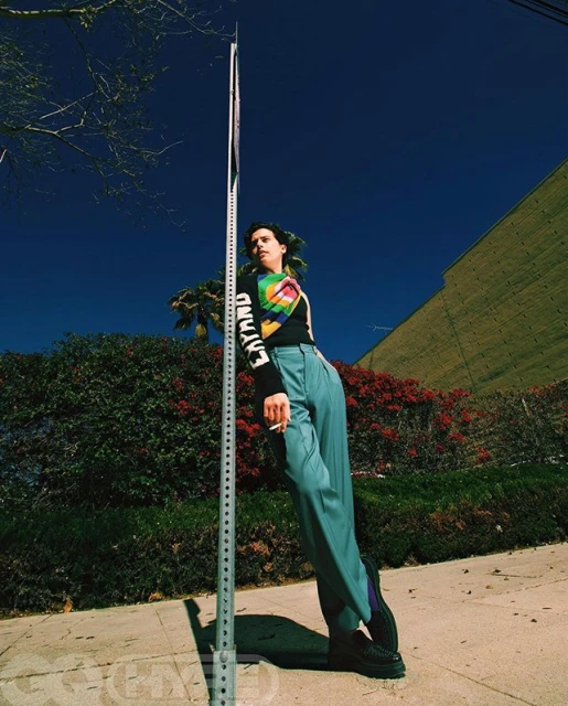 A young woman leans against a metal pole in a blue-toned fashion shot with greenery, a building, and bright blue sky visible.