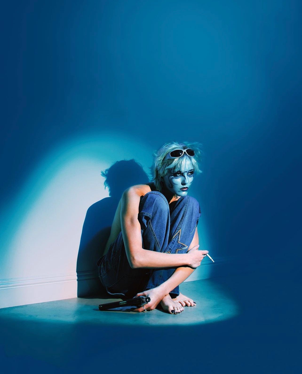 Woman with blue body paint sits on floor with cigarette in a photo studio. She wears sunglasses and jeans in blue light.