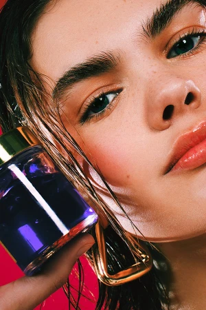 Close-up of a woman's face with wet hair, makeup, and statement earrings, holding a purple perfume bottle against a red backdrop.