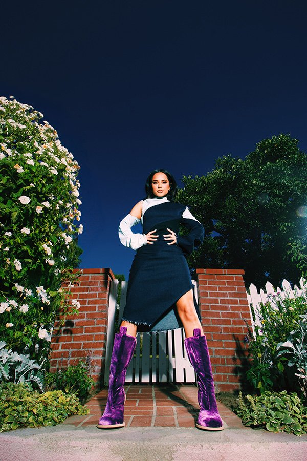 Woman in a black and white dress with purple velvet boots stands in front of a brick gate in a garden setting at dusk.