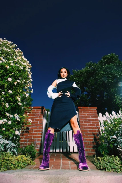 Woman in a black and white dress with purple velvet boots stands in front of a brick gate in a garden setting at dusk.