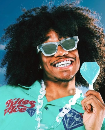 Man with afro, sunglasses, and turquoise shirt holds a diamond-shaped lollipop against a partly cloudy blue sky in a close-up studio shot.