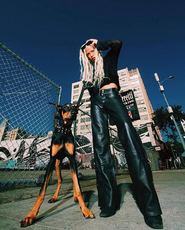 Fashion shot of a blonde-haired woman with a Doberman dog wearing a spiked collar on a city street under a blue sky.