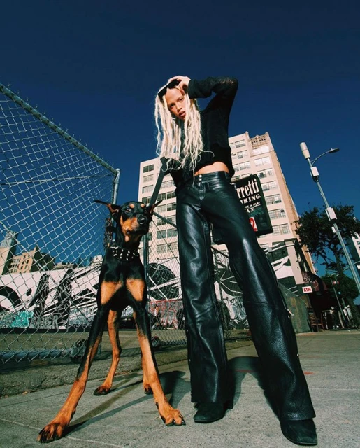 Fashion shot of a blonde-haired woman with a Doberman dog wearing a spiked collar on a city street under a blue sky.