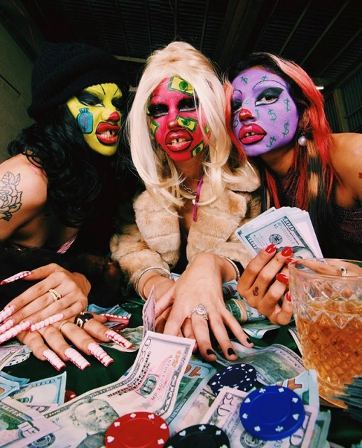 Three performers in colorful face paint pose behind a table piled with cash, casino chips, and a whiskey glass in an indoor setting.