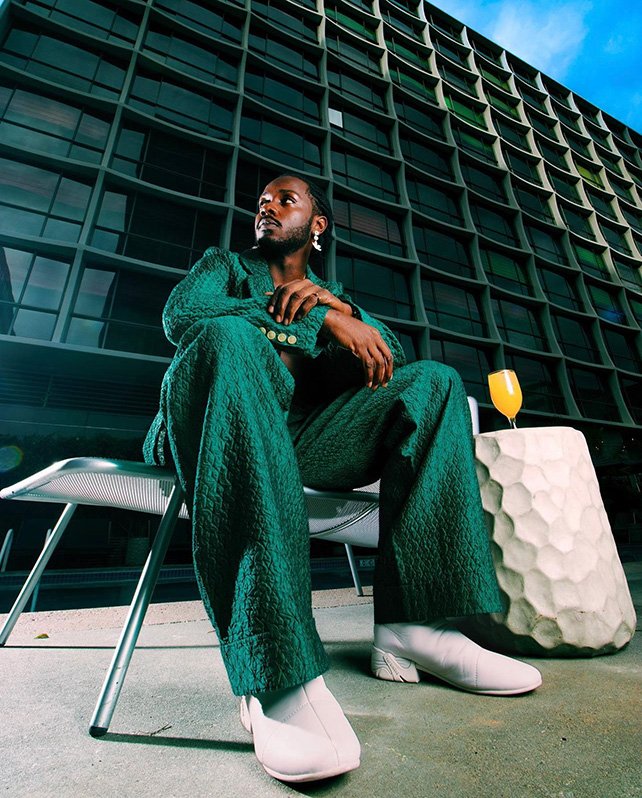 An African-American man in a green suit sits on a chair outdoors with a building and drink table behind him, shot at eye-level.