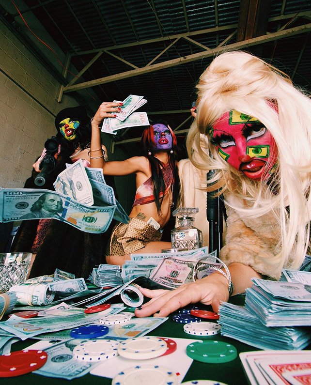 Three people with colorful face paint sit at a table covered in money and poker chips in an indoor setting, with an overhead view.