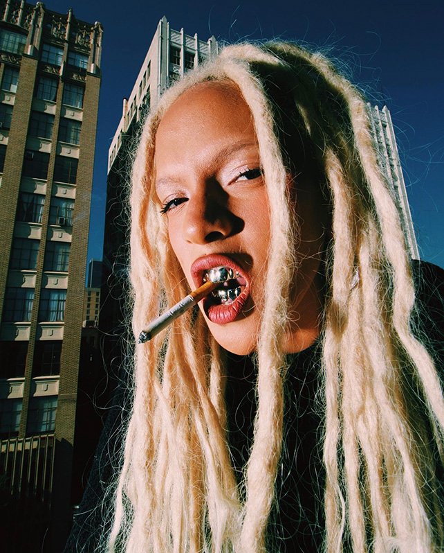 Close-up of a person with light skin, long blonde dreadlocks, grills, and a cigarette, set against a backdrop of tall buildings.