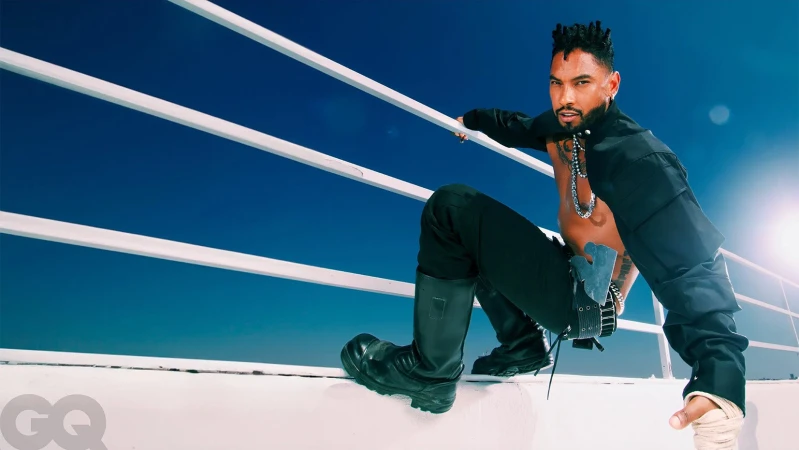 Black man wearing black trousers and tall boots crouching against a white railing with blue sky