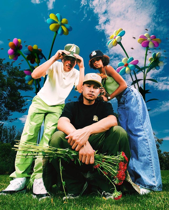 Three young adults pose outdoors in a field with oversized floral props under a sunny, partly cloudy sky, wearing streetwear.