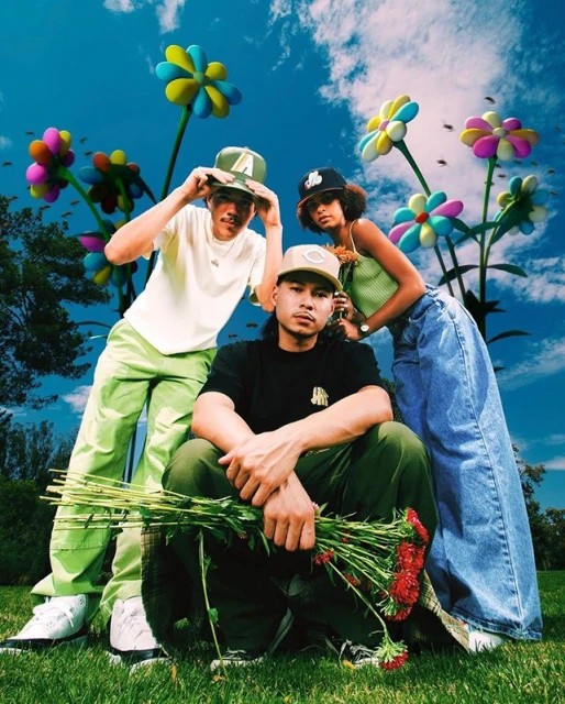 Three young adults pose outdoors in a field with oversized floral props under a sunny, partly cloudy sky, wearing streetwear.