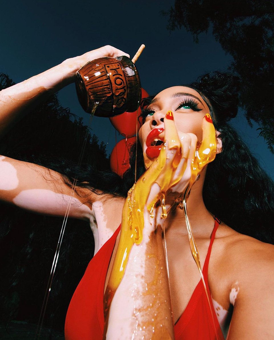 Woman pours honey on herself in a low-angle shot with a dark background, featuring a red top, dark hair, and a honey jar.