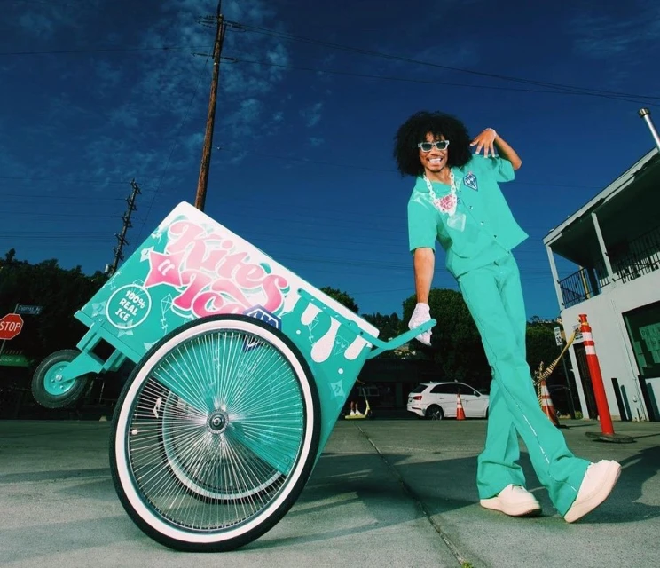 Man in coordinated turquoise outfit poses with matching vintage ice cream cart on city street under a clear blue sky, low angle.