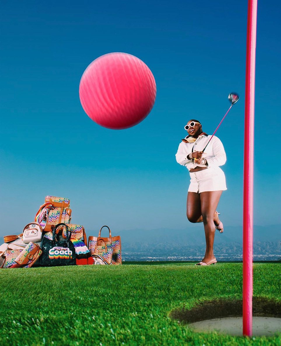 A fashionable Black woman prepares to tee off on a lush green golf course with designer bags and a giant pink golf ball.