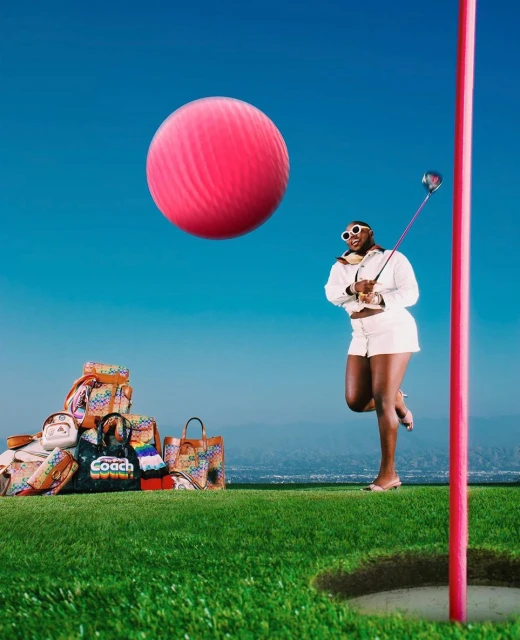 A fashionable Black woman prepares to tee off on a lush green golf course with designer bags and a giant pink golf ball.