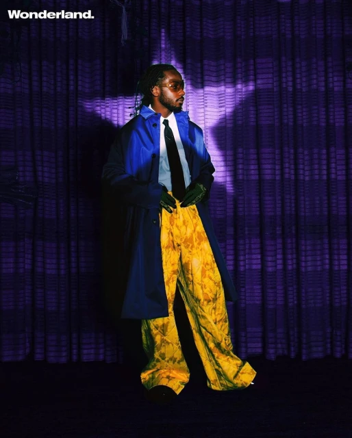 Full-length portrait of a Black male in a blue overcoat, yellow trousers, black tie and dark glasses against a purple curtain backdrop.