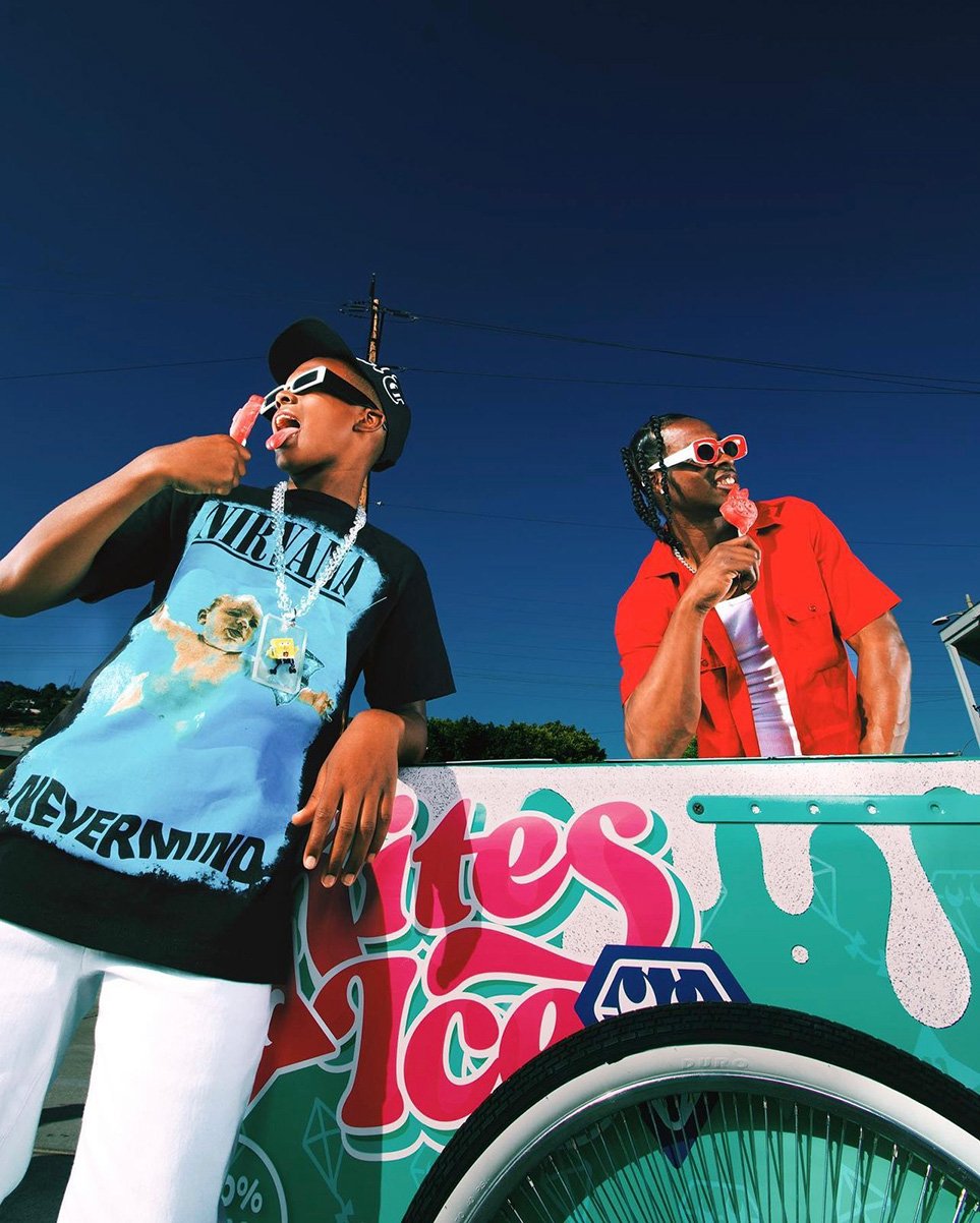 Two young men outdoors by ice cream cart, eating popsicles, wearing sunglasses and casual clothing against a blue sky.
