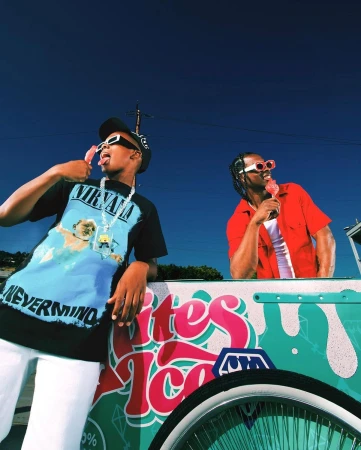 Two young men outdoors by ice cream cart, eating popsicles, wearing sunglasses and casual clothing against a blue sky.