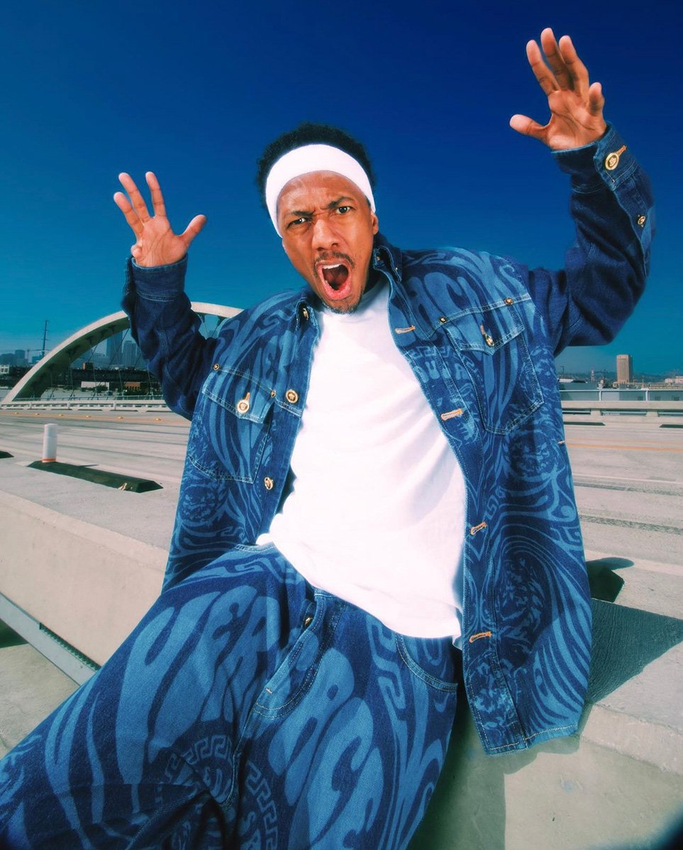 Man in patterned denim jacket and pants strikes a pose on a bridge against a blue sky with city skyline in the background.