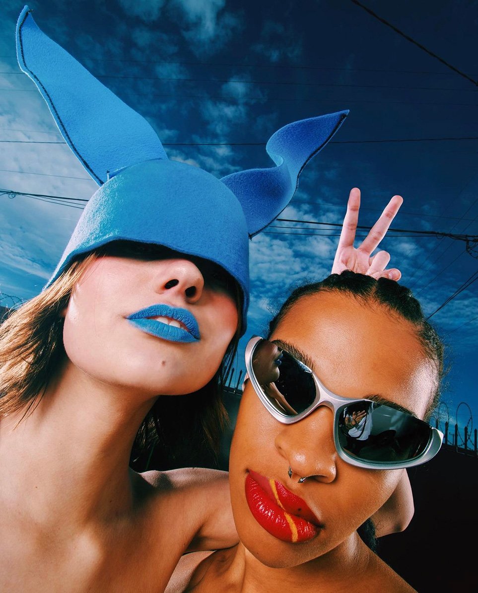 Two women pose in an outdoor setting, one with bright blue lipstick and a blue hat, the other with red lips and sunglasses.
