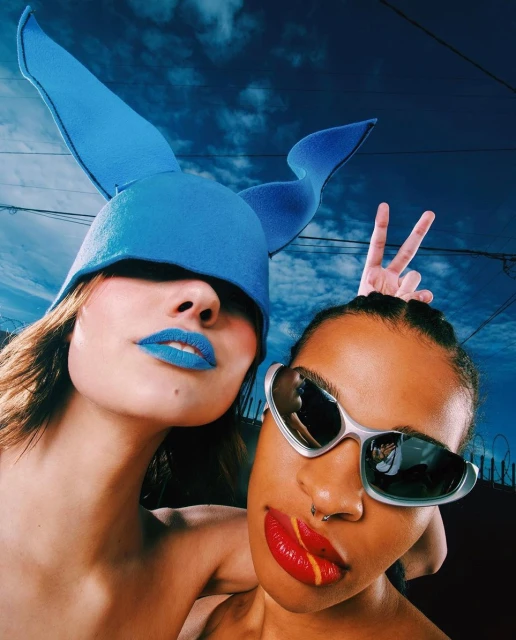 Two women pose in an outdoor setting, one with bright blue lipstick and a blue hat, the other with red lips and sunglasses.