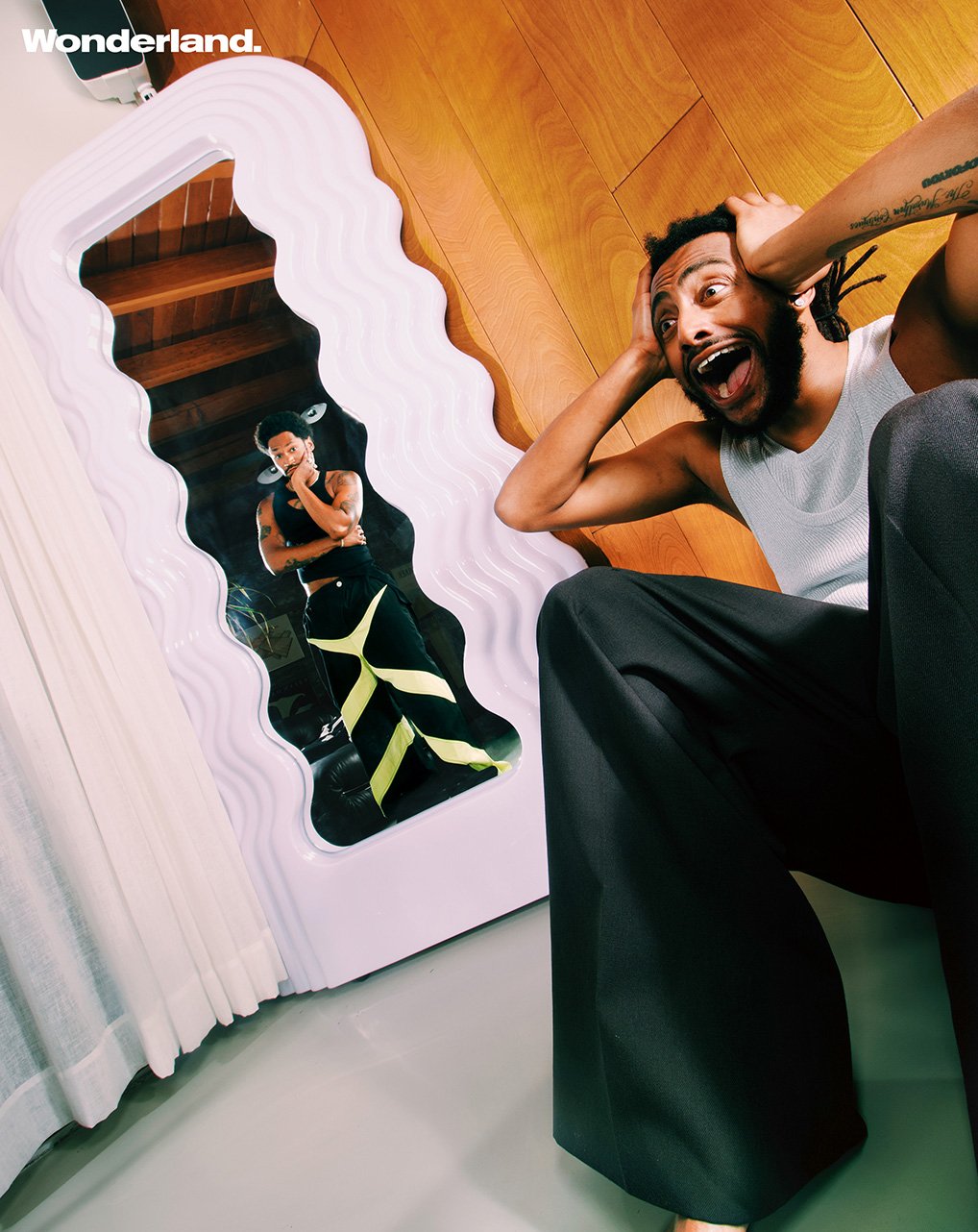 Man reacting near a mirror with another man's reflection, wearing a white tank top and black pants, in an indoor setting.