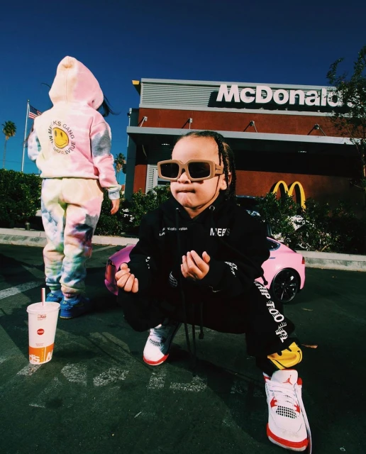 Two young children in trendy streetwear pose near a pink toy car and a McDonald's restaurant on a sunny day with blue sky.