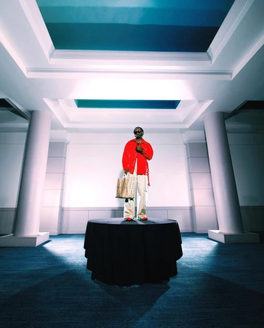Adult standing on a round table wearing a red jacket holding a tan patterned bag in an interior architectural space with columns.
