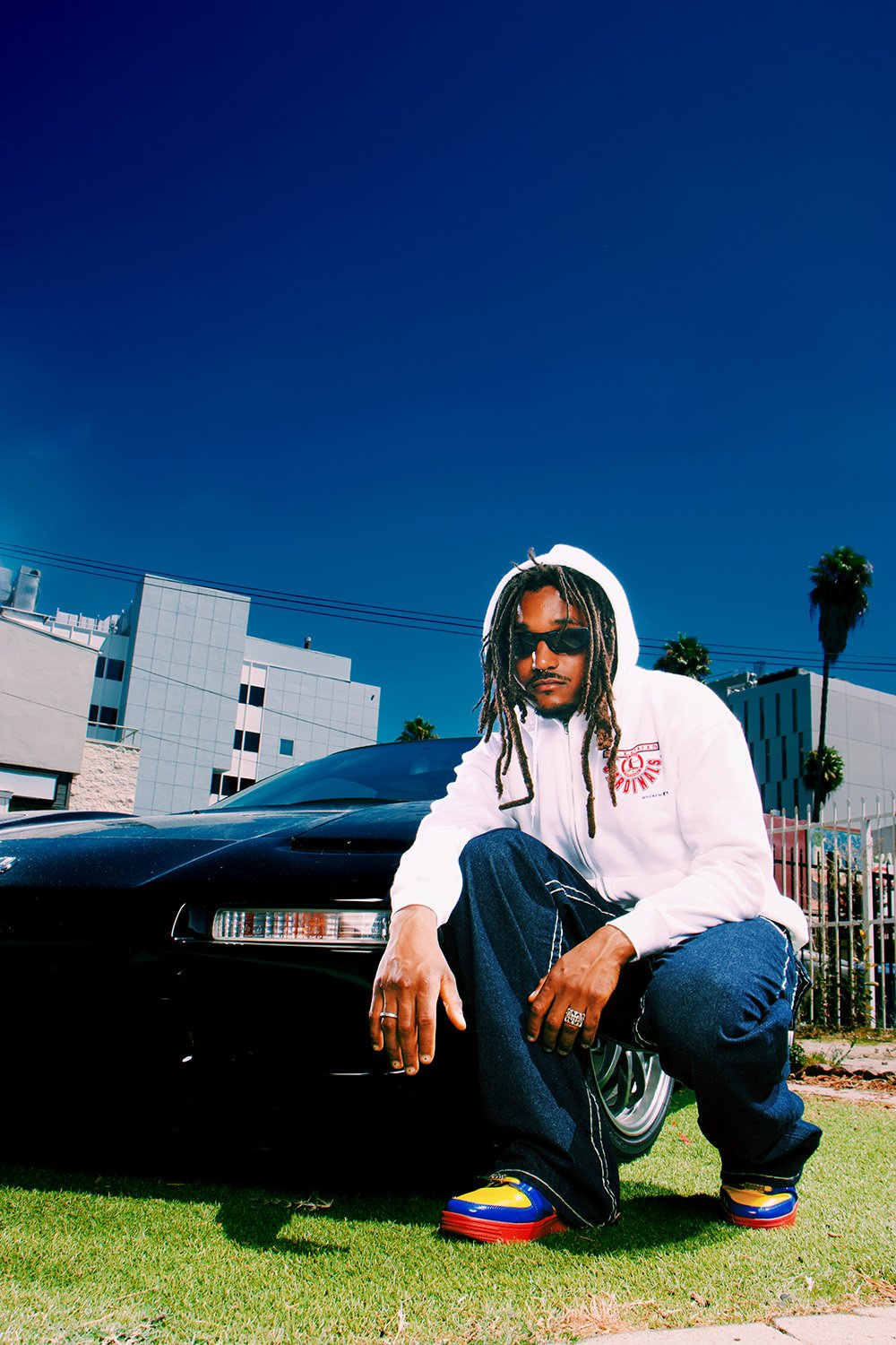 Adult male crouches next to a black car on a grassy lawn under a bright blue sky wearing a white hoodie, jeans, and colorful shoes.