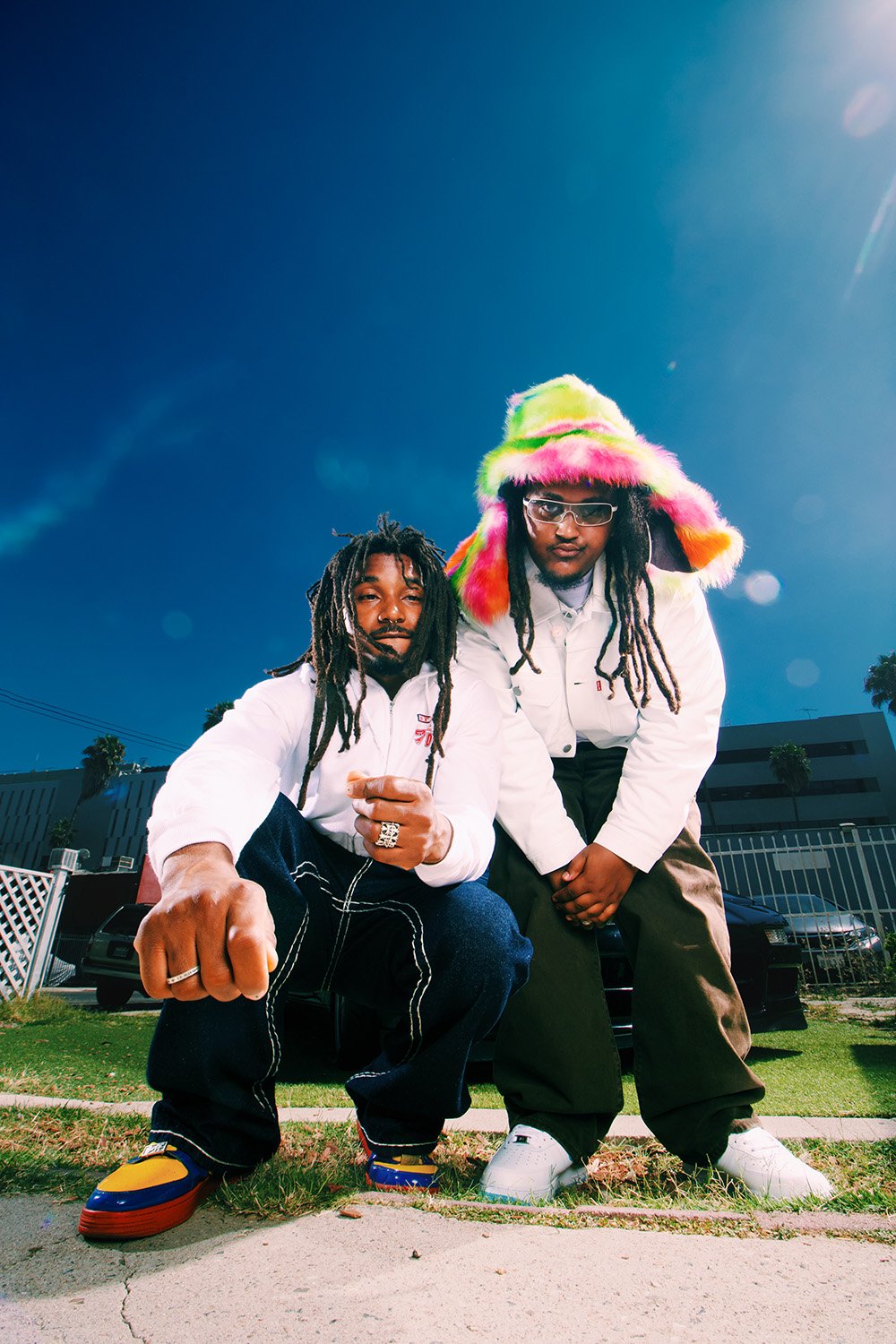 Two musicians squat in an urban setting with a car in the background, wearing streetwear, hats, and sneakers. The sky is blue.