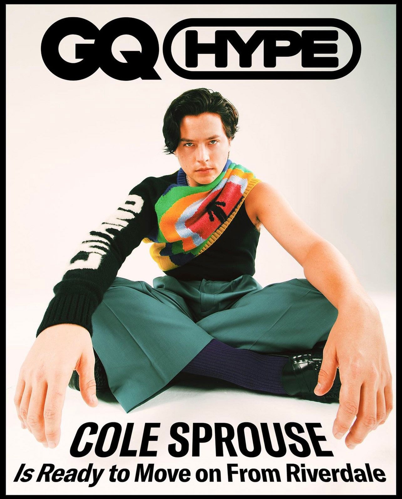 Cole Sprouse seated cross-legged on a white background wearing a black asymmetrical knit top with rainbow stripes and green trousers.