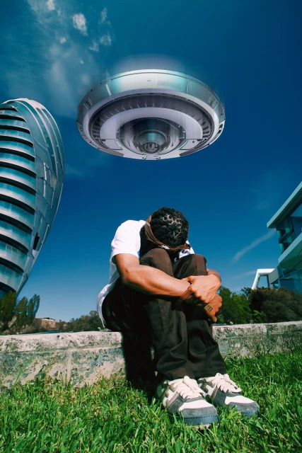 Man seated on a low wall with an apparent UFO hovering in a bright blue sky between modern buildings and green grass in a stylized composite image.