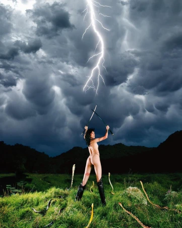 Woman in bikini and boots holding swords under dramatic thunderstorm in outdoor setting surrounded by faux snakes on the ground.