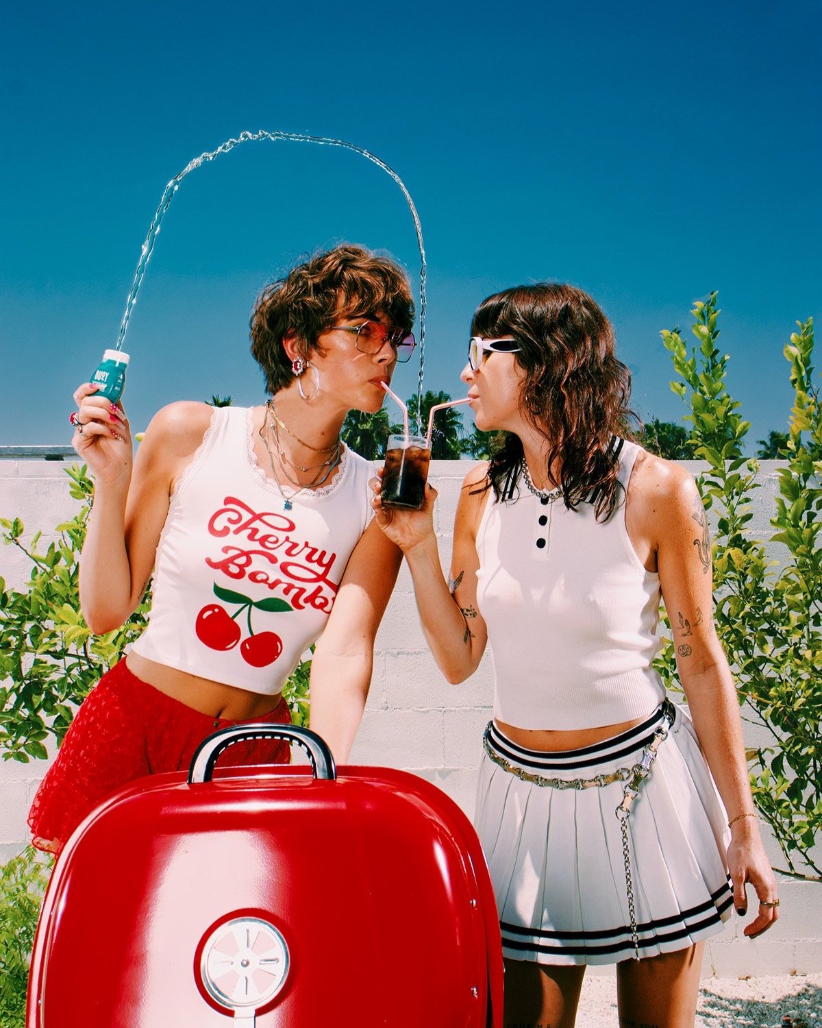 Two young women share a beverage outdoors on a sunny day, with a red grill in the foreground and blue sky in the background.