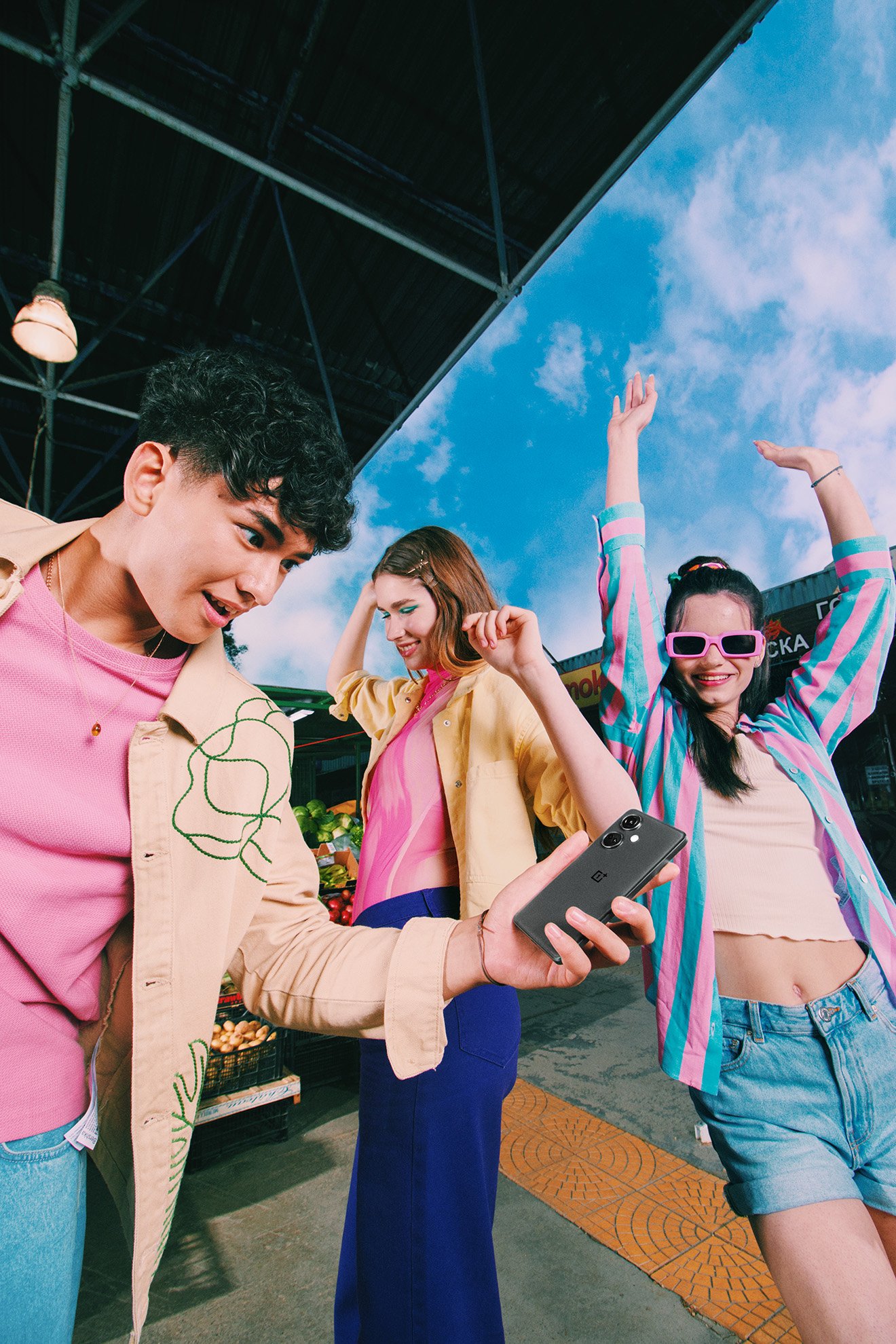 Three young adults pose with a mobile phone in an outdoor marketplace on a sunny day, showcasing modern casual fashion.
