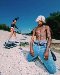 Shirtless tattooed man kneeling outdoors with a woman holding a roller behind him on a paved slope under blue sky