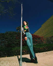 Low-angle shot of a man leaning against a metal street sign post wearing teal trousers and a black top