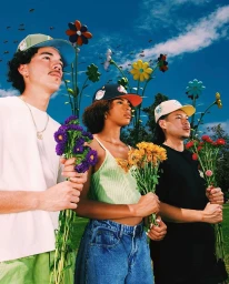 Three young models wearing baseball caps holding bouquets of flowers against a clear sky with giant floating flowers.