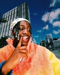 Low angle portrait of a smiling Black man with braided hair and beads wearing a colorful sherpa fleece jacket against a city backdrop.