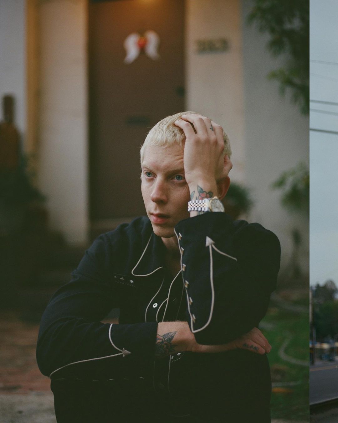 Portrait of a blond male with black western-style shirt sitting outside a white house on a city street, wearing a watch.