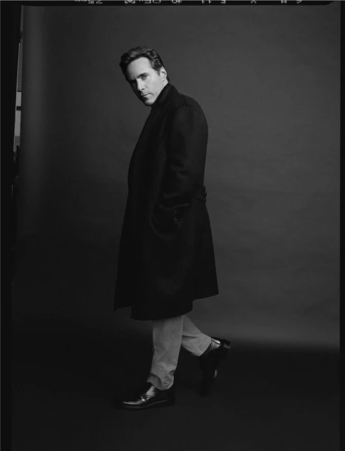 Monochrome portrait of a man in a long black coat walking in a dark studio. He has dark hair and is wearing light-colored trousers.