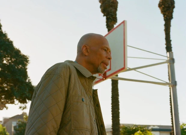 Profile shot of an older man with a bald head, short beard, and tan quilted jacket standing near a basketball hoop on a sunny day.