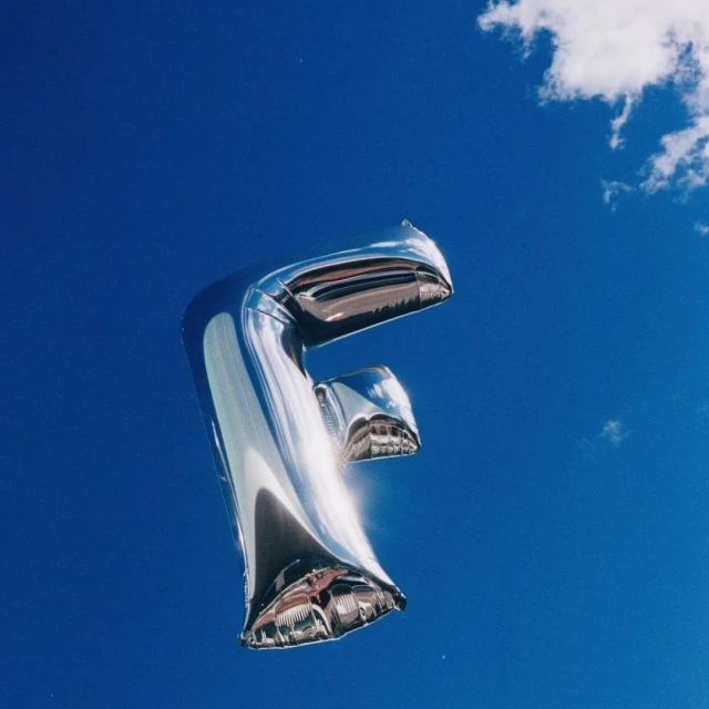 Silver letter F balloon against a bright blue sky with a few small clouds, creating a simple, celebratory, and visually striking composition.
